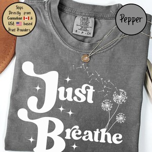 May include: Grey t-shirt with the words "Just Breathe" in white script and a dandelion design. The shirt has a crew neck and short sleeves. A silver necklace with a small pendant is visible.