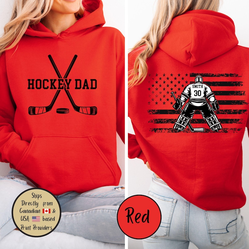 Puede incluir: Sudadera con capucha roja con "HOCKEY DAD" en letras negras, palos de hockey cruzados y un disco. La parte trasera presenta un gr&aacute;fico de portero de hockey con el nombre "SMITH" y el n&uacute;mero "30" sobre un dise&ntilde;o de bandera estadounidense en blanco y negro.