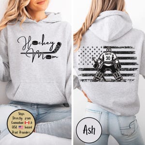 May include: Light gray hoodie with a front graphic that reads "Hockey Mom" with a hockey stick and puck. The back features a hockey goalie in front of a distressed American flag design. The goalie's jersey has the number 30 and the name "Smith".
