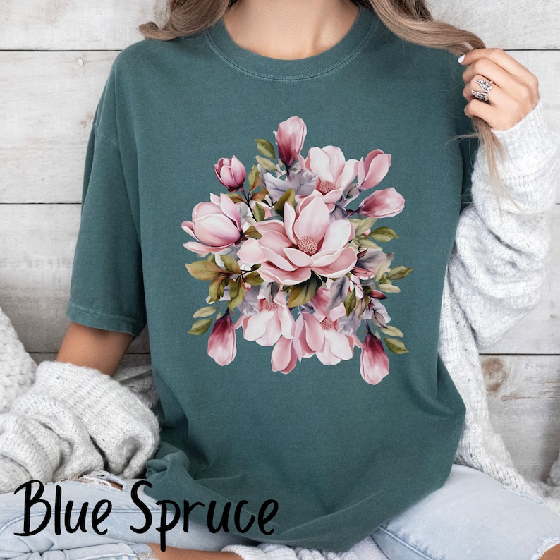 Peut inclure: Un t-shirt bleu sarcelle avec un motif floral compos&eacute; de fleurs de magnolia roses et de feuilles vertes.