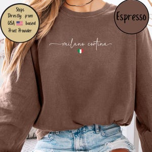 May include: A brown sweatshirt with the words "milano cortina" in a cursive font, and a small Italian flag graphic. The sweatshirt has long sleeves and is made of a soft material. The text "Espresso" is in a brown circle.