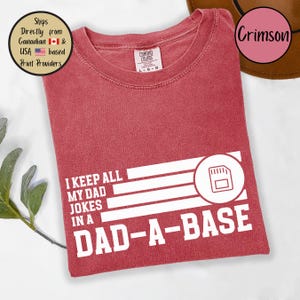 May include: Crimson-colored t-shirt with the text "I KEEP ALL MY DAD JOKES IN A DAD-A-BASE" in white. The design includes a graphic of a floppy disk. The shirt is from Comfort Colors.