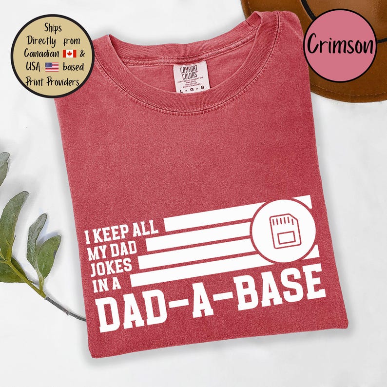 May include: Crimson-colored t-shirt with the text "I KEEP ALL MY DAD JOKES IN A DAD-A-BASE" in white. The design includes a graphic of a floppy disk. The shirt is from Comfort Colors.