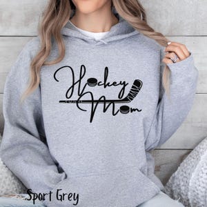 Op de afbeelding: Een grijs hoodie met de tekst "Hockey Mom" in zwarte letters. Een hockeystick en een puck zijn opgenomen in het ontwerp.