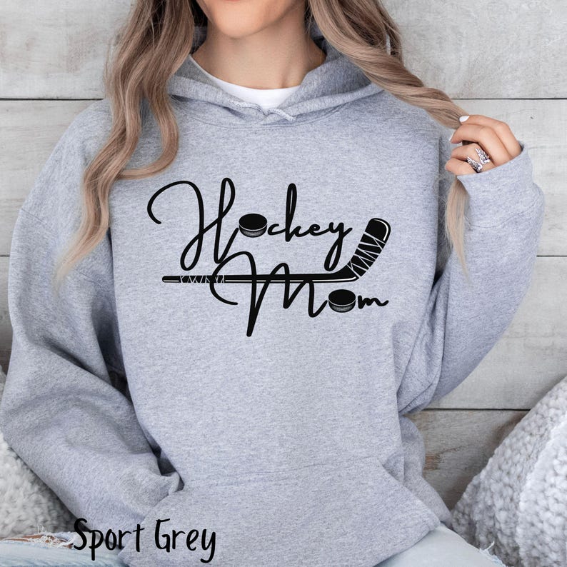 Op de afbeelding: Een grijs hoodie met de tekst "Hockey Mom" in zwarte letters. Een hockeystick en een puck zijn opgenomen in het ontwerp.