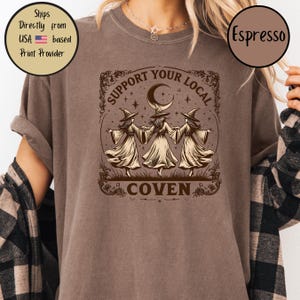May include: Espresso-colored t-shirt featuring a graphic of three witches holding hands under a crescent moon and stars, with the text "SUPPORT YOUR LOCAL COVEN". The design is in a vintage style.