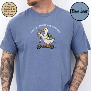 May include: Blue short-sleeved t-shirt with the text "Off to Commit Dad Foolery." The shirt features a cartoon duck riding a scooter while holding a coffee cup. The duck wears a backpack. The shirt is blue.