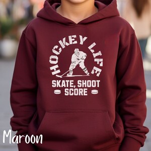 Op de afbeelding: Een maroon hoodie met een witte afbeelding van een hockeyer in een cirkel met de tekst "Hockey Life" en "Skate, Shoot Score" onder de cirkel.
