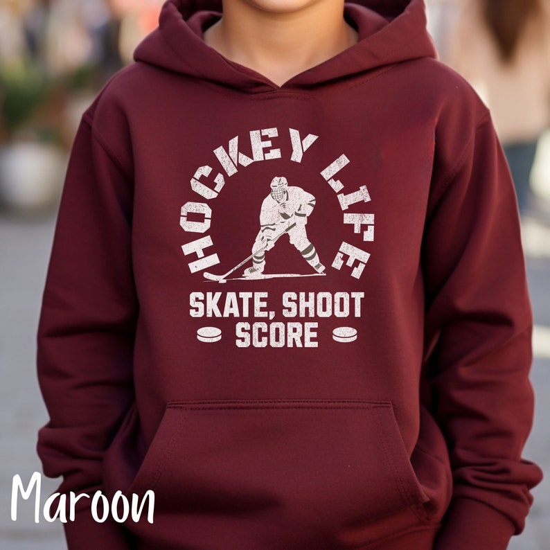 Op de afbeelding: Een maroon hoodie met een witte afbeelding van een hockeyer in een cirkel met de tekst "Hockey Life" en "Skate, Shoot Score" onder de cirkel.