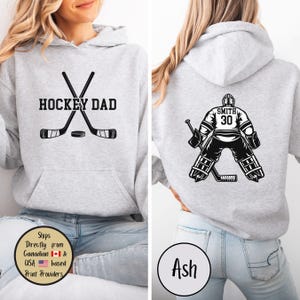 Puede incluir: Sudadera con capucha gris claro con un gr&aacute;fico frontal que dice "HOCKEY DAD" con palos de hockey cruzados y un disco. La parte trasera presenta un gr&aacute;fico de portero de hockey con el n&uacute;mero 30 y el nombre "SMITH".