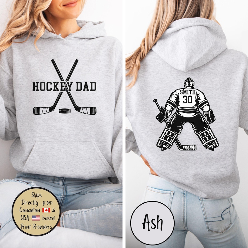 Puede incluir: Sudadera con capucha gris claro con un gr&aacute;fico frontal que dice "HOCKEY DAD" con palos de hockey cruzados y un disco. La parte trasera presenta un gr&aacute;fico de portero de hockey con el n&uacute;mero 30 y el nombre "SMITH".