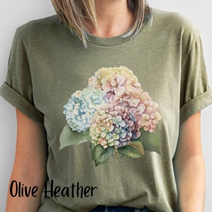 May include: A green t-shirt with a watercolor floral design featuring a bouquet of hydrangeas in shades of pink, blue, and white. The text "Olive Heather" is printed on the bottom of the shirt.