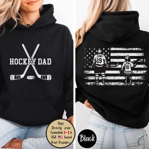 Puede incluir: Sudadera con capucha negra con el texto "HOCKEY DAD" y palos de hockey cruzados y un disco. La parte trasera presenta un dise&ntilde;o de bandera estadounidense con jugadores de hockey y el texto "KING 19" y "KING 91".