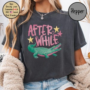 May include: Dark gray t-shirt with the text "AFTER WHILE" in pink and yellow stars. A green alligator with pink accents is below the text. The shirt is a casual style.
