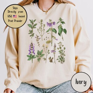 May include: Ivory-colored sweatshirt featuring a botanical print of various wildflowers in shades of green, purple, and yellow. The design includes detailed illustrations of leaves and stems. The text "Ivory" is printed on the bottom right.