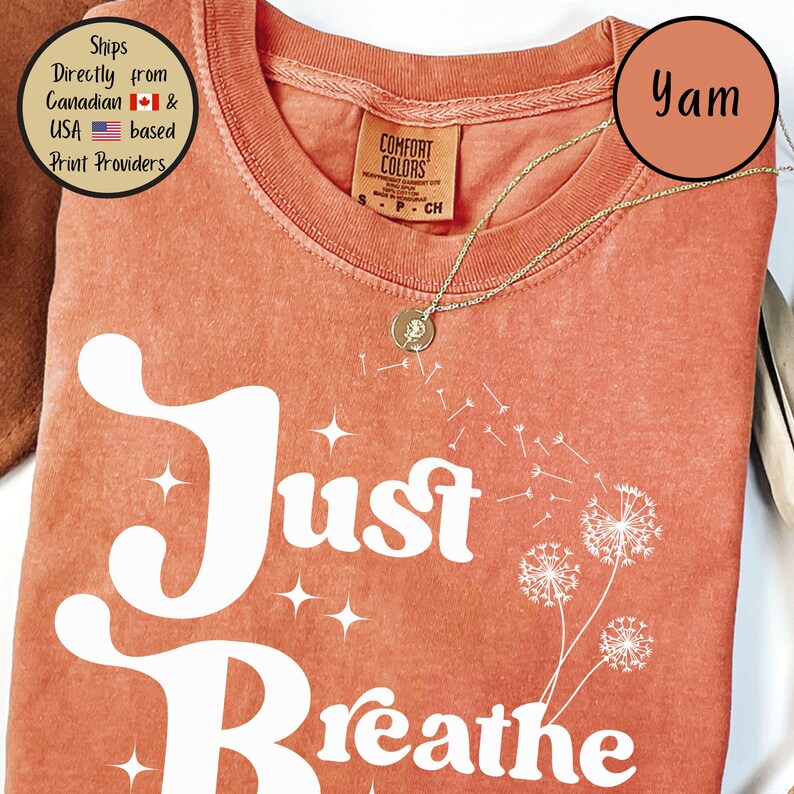 May include: A coral-coloured t-shirt with the words "Just Breathe" in white script and a dandelion graphic. A gold necklace with a pendant rests on the shirt. The shirt has a Comfort Colors tag.