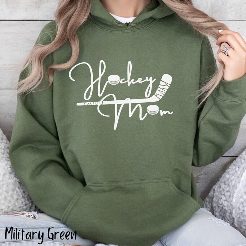 Op de afbeelding: Een groene hoodie met de tekst "Hockey Mom" in witte letters. Het ontwerp bevat een hockeystick en een puck.
