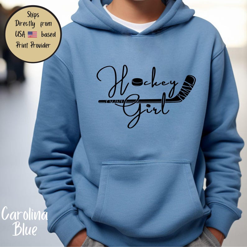May include: Carolina blue hoodie with a front pocket. The hoodie features the words "Hockey Girl" in a script font, along with a hockey puck and stick graphic in black. The hoodie is made of a soft material.