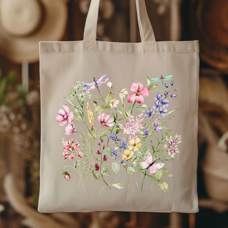May include: A beige tote bag with a floral design featuring pink, purple, yellow, and white flowers, green leaves, and dragonflies. The design is centered on the bag.