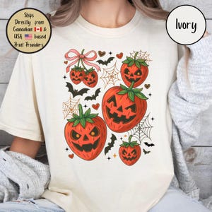 May include: Ivory-colored t-shirt featuring a Halloween-themed graphic. The design includes orange jack-o'-lanterns, bats, spiderwebs, and hearts. The shirt has a crew neck and short sleeves. The text "Ivory" is in a circle.
