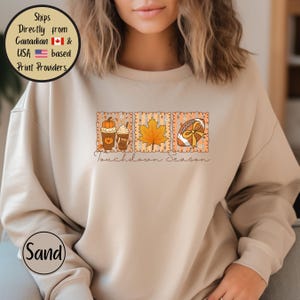 May include: Sand-colored sweatshirt featuring a fall-themed graphic. The graphic includes two coffee cups with pumpkin and whipped cream, a maple leaf, and a football with a bow. The text "Touchdown season" is written below the graphic.