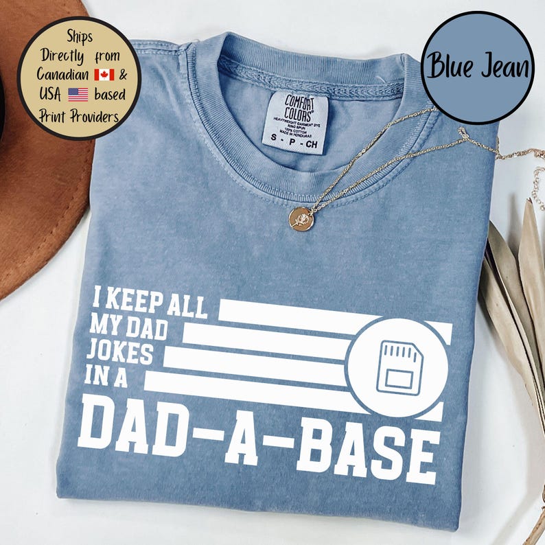 May include: Blue jean colored t-shirt with the text "I KEEP ALL MY DAD JOKES IN A DAD-A-BASE" in white. The graphic includes a stylized data storage icon. A gold necklace is draped on the shirt.