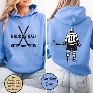Puede incluir: Sudadera con capucha azul Carolina con "HOCKEY DAD" impreso en la parte delantera, con palos de hockey cruzados y un disco. La parte trasera muestra un gr&aacute;fico de jugador de hockey con el nombre "SCHUMACHER" y el n&uacute;mero "11".