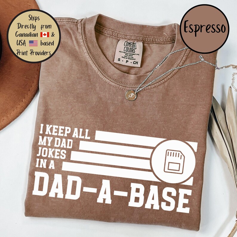 May include: Espresso-colored t-shirt with the text "I KEEP ALL MY DAD JOKES IN A DAD-A-BASE" in white. The shirt has a crew neck and short sleeves. A silver necklace with a pendant is visible.