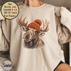 May include: A sand-coloured crewneck sweatshirt featuring a graphic illustration of a brown moose wearing a burnt orange knit beanie with a pom-pom. The moose is centred on the chest, showcasing detailed fur texture and large antlers.