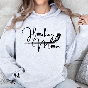 Op de afbeelding: Grijs hoodie met de tekst "Hockey Mom" in zwarte schrijfletters met een grafische afbeelding van een hockeystick en een puck.