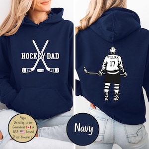 Puede incluir: Sudadera con capucha azul marino con gr&aacute;ficos blancos. La parte delantera presenta "HOCKEY DAD" con palos de hockey cruzados y un disco. La parte trasera muestra un jugador de hockey con el nombre "WILLIAM" y el n&uacute;mero "17".