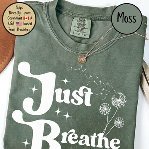 May include: A moss green t-shirt with the words "Just Breathe" in white script. The shirt features a dandelion graphic and a gold necklace. The shirt is made by Comfort Colors.