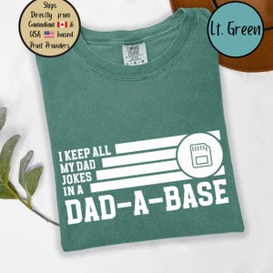 May include: A light green t-shirt with the text "I KEEP ALL MY DAD JOKES IN A DAD-A-BASE" in white. The design includes a graphic of a memory card. The shirt is made of Comfort Colors material.