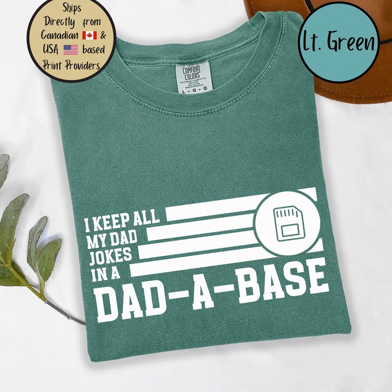 May include: A light green t-shirt with the text "I KEEP ALL MY DAD JOKES IN A DAD-A-BASE" in white. The design includes a graphic of a memory card. The shirt is made of Comfort Colors material.