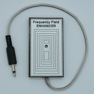 Könnte beinhalten: Ein rechteckiges Gerät mit dem Text "Frequency Field ENHANCER" auf einer weißen Oberfläche. Das Gerät hat schwarze konzentrische Rechtecke und einen kleinen silbernen Knopf. Ein schwarzer Stecker und ein graues Kabel sind angebracht.