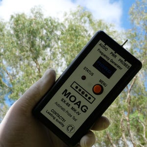 May include: A black and white electronic device with the text "MOAG SX-88 MK-IV Automatic Probe Test" and "Frequency Generator". The device has a digital display and several buttons. The background is blurred green foliage.