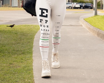 May include: A woman wearing white leggings with a black eye chart print and a white sweater. She is holding a cup of coffee and walking down a sidewalk.