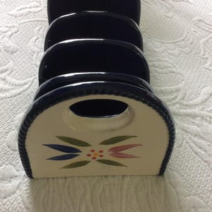 May include: A ceramic letter holder with a navy blue finish and a cream-colored front. The front features a floral design in blue, green, and pink. The holder has a handle at the top and is designed to hold mail or documents.
