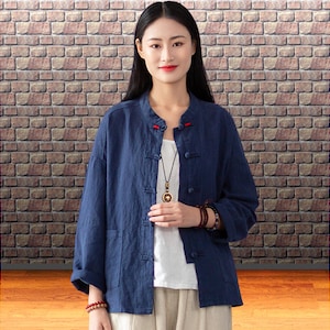 May include: A navy blue linen jacket with a mandarin collar and three-quarter sleeves. The jacket has a button-down front, two front pockets, and red accents at the collar. The model is wearing a white top and beige pants.