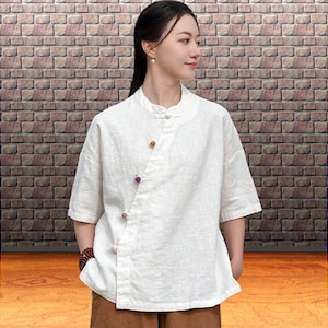 May include: A white linen shirt with a mandarin collar and short sleeves. The shirt has an asymmetrical button closure with three decorative buttons in shades of brown and purple. The model is wearing brown trousers and a beaded bracelet.