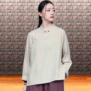 Könnte beinhalten: Ein beiges Langarmshirt mit Stehkragen und roten Knöpfen. Das Shirt hat eine lockere Passform und wird mit einer dunkelvioletten Hose kombiniert. Der Hintergrund zeigt eine Backsteinmauer.