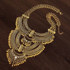 Make a bold impact with this antique gold bib necklace, featuring intricate engravings and resin stones designed for high-drama festival looks. Whether you are channeling a desert queen or elevating a bohemian outfit, this layered piece sits perfectly against the collarbone to command attention. Inspired by traditional nomadic adornments, this necklace uses weightier textures and a multi-tiered design to create movement and sound as you move.