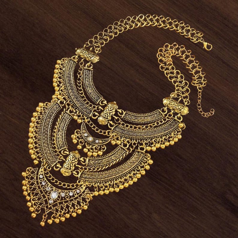 Make a bold impact with this antique gold bib necklace, featuring intricate engravings and resin stones designed for high-drama festival looks. Whether you are channeling a desert queen or elevating a bohemian outfit, this layered piece sits perfectly against the collarbone to command attention. Inspired by traditional nomadic adornments, this necklace uses weightier textures and a multi-tiered design to create movement and sound as you move.