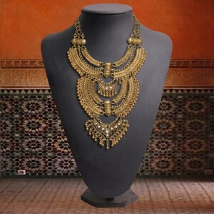 Make a bold impact with this antique gold bib necklace, featuring intricate engravings and resin stones designed for high-drama festival looks. Whether you are channeling a desert queen or elevating a bohemian outfit, this layered piece sits perfectly against the collarbone to command attention. Inspired by traditional nomadic adornments, this necklace uses weightier textures and a multi-tiered design to create movement and sound as you move.
