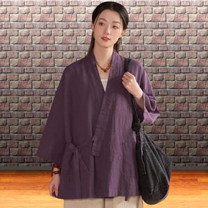 May include: A plum-colored kimono-style jacket with a tie closure and pockets. The jacket is worn over a white top and paired with beige pants. A black tote bag is carried over the shoulder. The background features a brick wall.