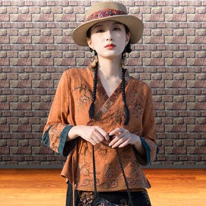 Puede incluir: Una blusa envolvente naranja con diseños florales bordados y detalles turquesa. La blusa se combina con un sombrero de paja con una banda estampada. El conjunto es una mezcla de estilos tradicionales y contemporáneos.