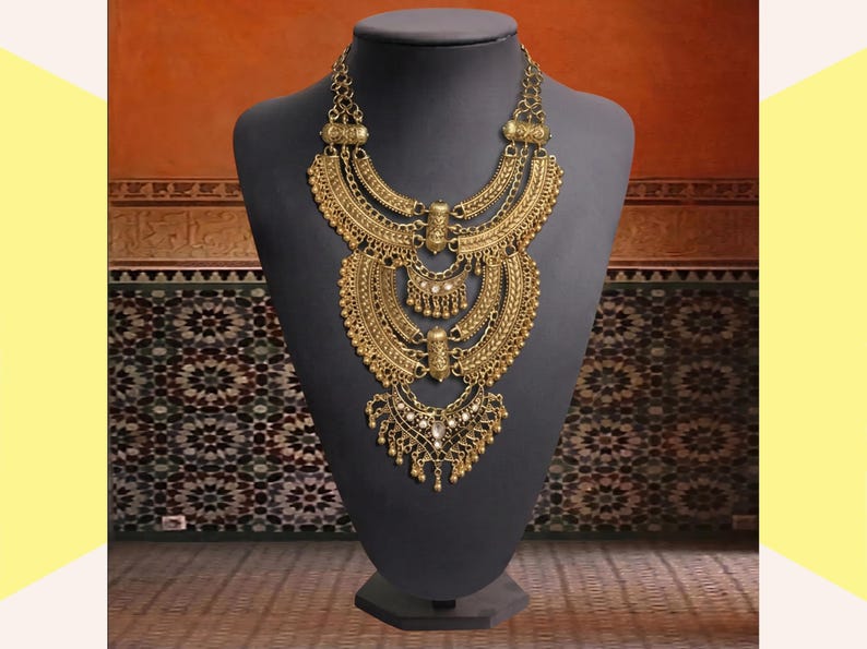 Make a bold impact with this antique gold bib necklace, featuring intricate engravings and resin stones designed for high-drama festival looks. Whether you are channeling a desert queen or elevating a bohemian outfit, this layered piece sits perfectly against the collarbone to command attention. Inspired by traditional nomadic adornments, this necklace uses weightier textures and a multi-tiered design to create movement and sound as you move.