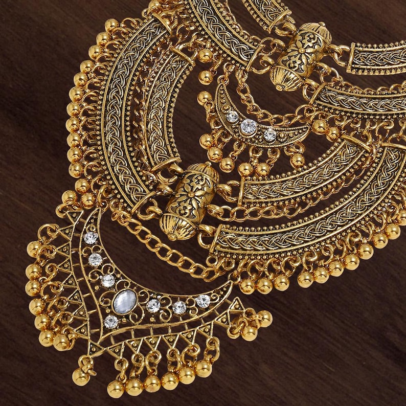 Make a bold impact with this antique gold bib necklace, featuring intricate engravings and resin stones designed for high-drama festival looks. Whether you are channeling a desert queen or elevating a bohemian outfit, this layered piece sits perfectly against the collarbone to command attention. Inspired by traditional nomadic adornments, this necklace uses weightier textures and a multi-tiered design to create movement and sound as you move.