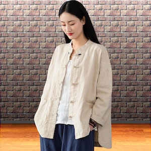 Peut inclure: Veste en lin beige avec un col mandarin et des fermetures à bascule en bois. La veste est portée sur un haut blanc et associée à un pantalon bleu marine. Le fond présente un mur de briques.