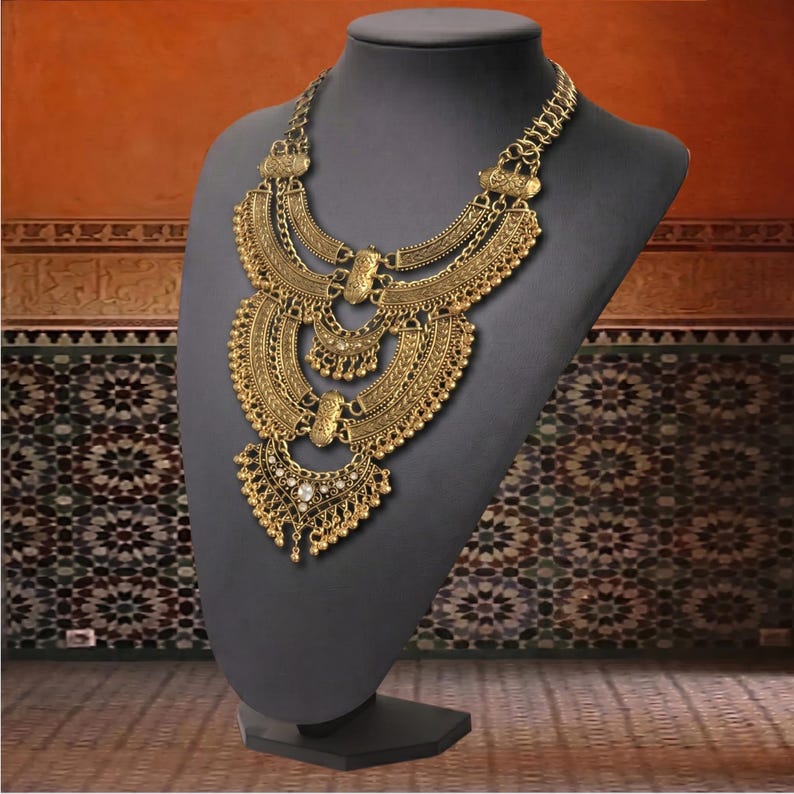 Make a bold impact with this antique gold bib necklace, featuring intricate engravings and resin stones designed for high-drama festival looks. Whether you are channeling a desert queen or elevating a bohemian outfit, this layered piece sits perfectly against the collarbone to command attention. Inspired by traditional nomadic adornments, this necklace uses weightier textures and a multi-tiered design to create movement and sound as you move.